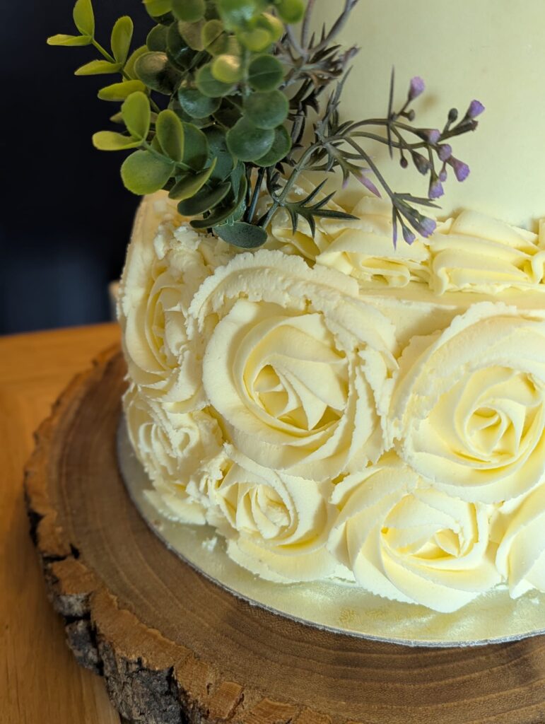 weddingpr2 Image shows a two-tier white cake on a log slice, decorated with buttercream roses and heather and eucalyptus.